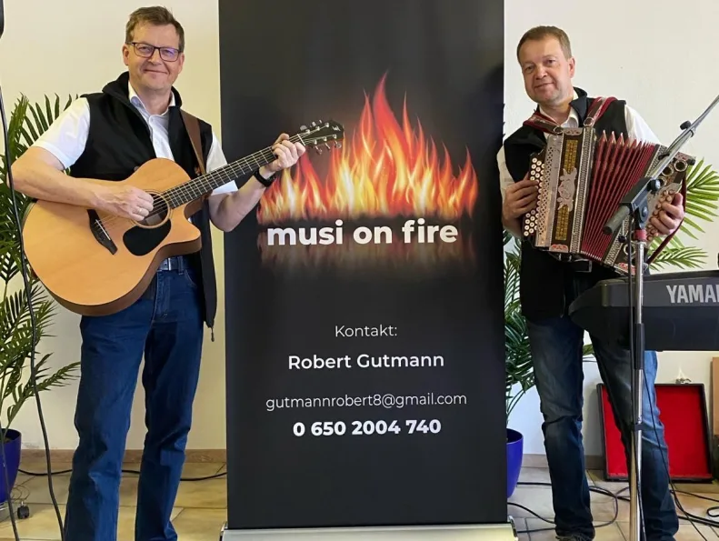 Foto: musi on fire Die beiden Musiker von musi on fire neben ihrem Roll-up mit dem Flammen-Logo – links mit Gitarre, rechts mit Steirischer Harmonika