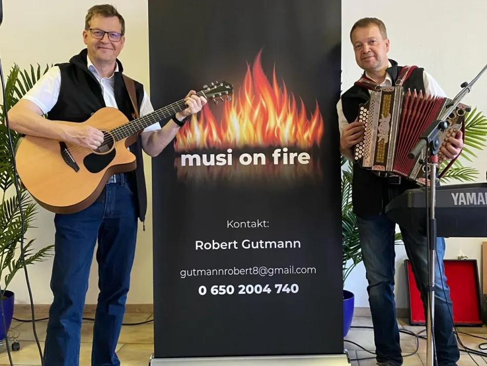 Foto: musi on fire Die beiden Musiker von musi on fire neben ihrem Roll-up mit dem Flammen-Logo – links mit Gitarre, rechts mit Steirischer Harmonika