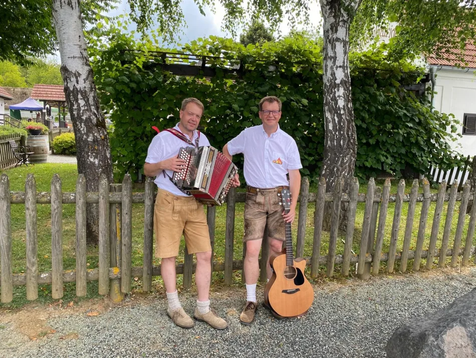 Foto: musi on fire Die beiden Musiker von musi on fire in Lederhose und weißem Hemd vor einem Holzzaun unter Birken – einer mit Steirischer Harmonika, einer mit Gitarre